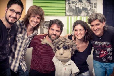 Group of five adults smiling around a sunglasses-wearing puppet in a casual studio setting, uskovat etta 13