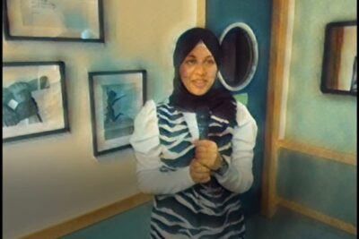 Woman wearing a hijab and patterned dress standing by framed artwork ready to uttrycka asikter