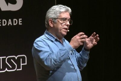 Man with gray hair and glasses speaking on stage with a headset microphone and gesturing while referencing vacinas sao.