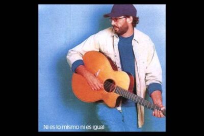 Guitarist in a cap and glasses holding an acoustic guitar against a blue background, a scene that suggests it may be vale pena.