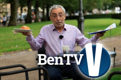 Older man at a park café table holding a bun on a plate and papers with a visible studio logo, vastainen keskustelu 1