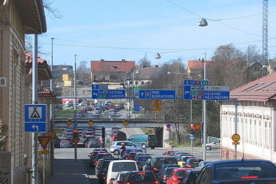 Congested urban street with rows of cars and overhead blue directional signs indicating vastakkaiseen suuntaan.