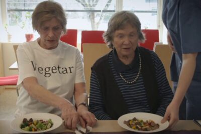 Two elderly women at a table with plates of mixed vegetables and a caregiver serving food, highlighting vegansk kosthold 1.