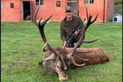 Hunter kneeling behind a large trophy stag on grass beside a red brick barn after a successful verdens jagt.