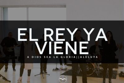 Band performing indoors with guitars, drums and vocalists beneath the Spanish headline 'El Rey Ya Viene' — vida real moment