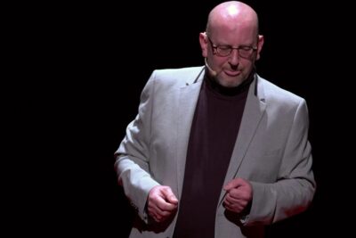 Bald presenter with glasses and headset microphone on a dark stage, ideal for vie virus related content.