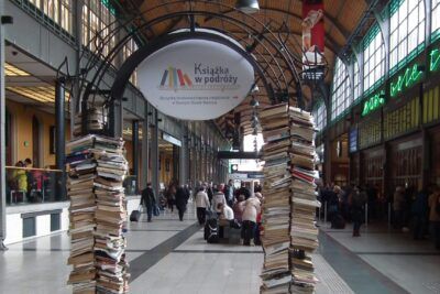 Tall columns of stacked books on wheeled carts in a busy train station concourse with arched ceiling and commuters, vigtigt at 11