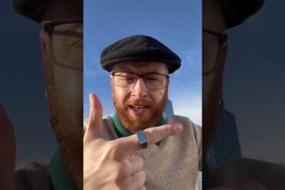 Bearded man with glasses and a flat cap pointing at the camera against a blue sky, viktig a 104.