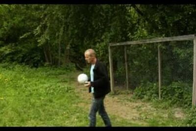 Adult man holding a white soccer ball beside a chain-link fence and trees, viktig a 168.