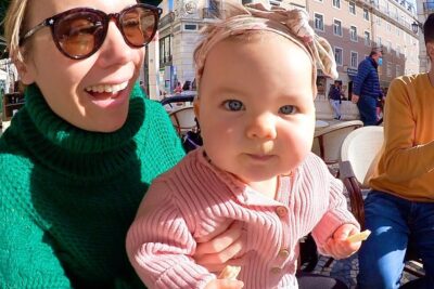 Smiling woman holding a baby in a pink sweater at an outdoor café in bright sunlight, a candid viktig a 7 family scene.