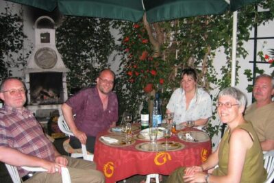 Group of five people seated around a red-covered garden table under an umbrella, surrounded by greenery where viljelykasvit voivat flourish.