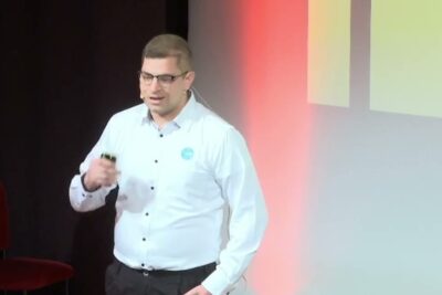 Man in a white shirt and glasses speaking on stage with a headset microphone advocating for vittime bullismo.