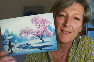 Woman holding a postcard-sized blue and pink cherry blossom winter painting with a small figure and snowy stream, vivere dellimmagine 1.