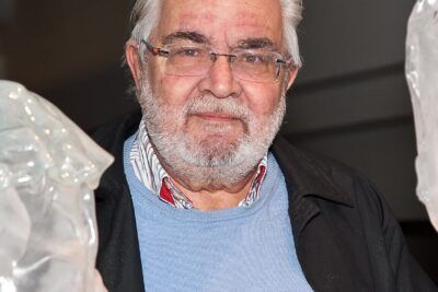 Elderly man with white beard and glasses wearing a light blue sweater and dark jacket, voi auttaa