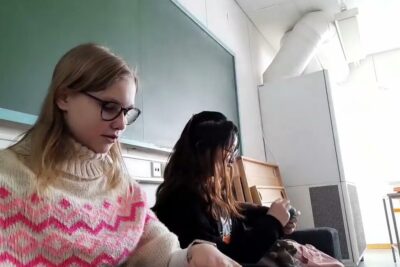 Two people seated in a classroom near a chalkboard, one wearing a patterned sweater and glasses, voi johtaa 12