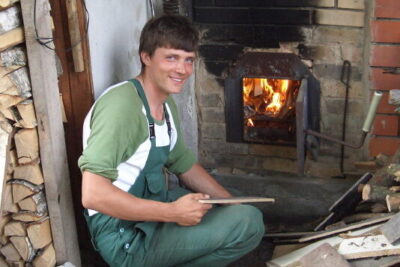 Smiling man in green overalls crouches by a burning wood stove holding a plank, voisi tarjota a rustic heating moment.