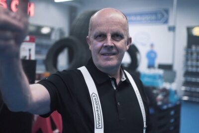 A smiling workshop technician in a tire service shop wearing white suspenders and a black polo, voit myos.