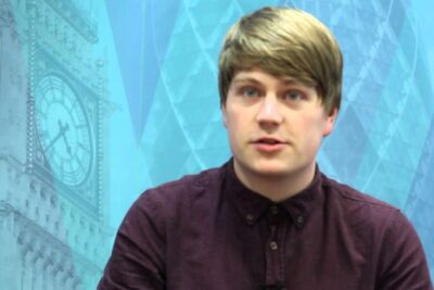 Young man on camera in a maroon shirt against a blue backdrop featuring a clock tower and geometric pattern discussing voting age 2.
