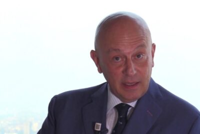 Bald professional in a dark suit speaking with a lapel microphone against a bright city backdrop about voto elettronico