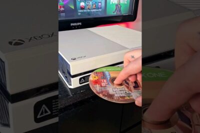 White xbox one 10 console with Grand Theft Auto V disc being inserted - sleek gaming setup on a black desk with a Philips monitor showing game thumbnails.
