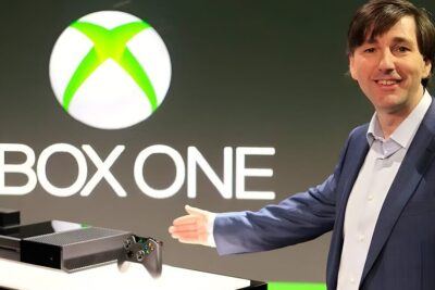 Presenter pointing to an xbox one 11 console and controller in front of a large Xbox One logo