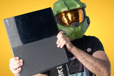 Person wearing a green Master Chief helmet holding an xbox one 9 console against a yellow background.