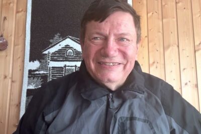Smiling man in a jacket against wooden-paneled wall, ydinaseiden kayton 1