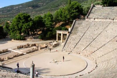 Wide view of a sunlit ancient amphitheater with circular orchestra and stepped stone seating, zeusin rooli 1.