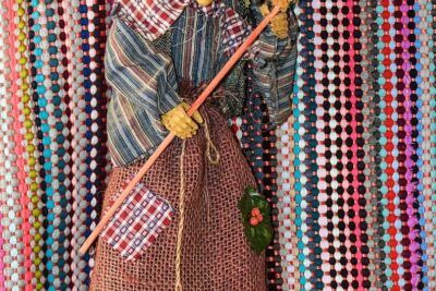 Handcrafted witch doll holding a broom against a colorful woven background, zombit ovat