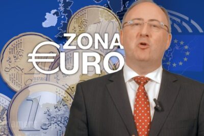 A suited presenter speaking in front of oversized euro coins and a map highlighting the zona euro