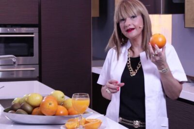 A woman in a modern kitchen holds an orange beside a glass of zumo naranja 3 on the counter.